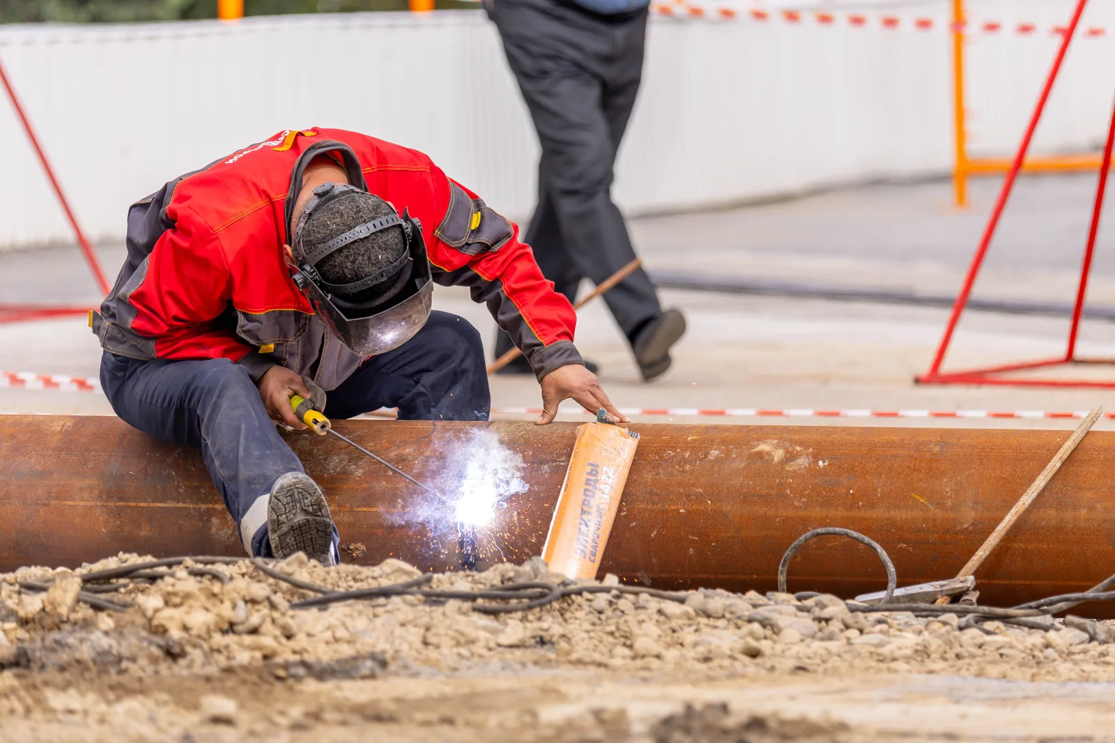 A worker in protective gear welding a joint on a large pipe laid in an open trench
