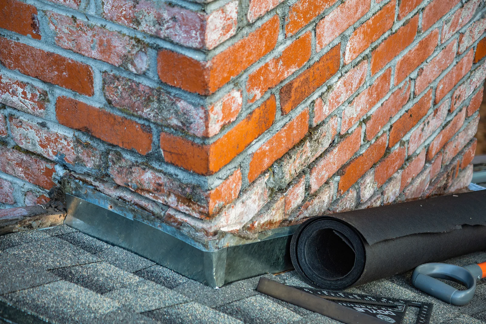 Metal step flashing set into the brickwork where a chimney meets an asphalt shingle roof, with a roll of membrane and a knife left on the slope.