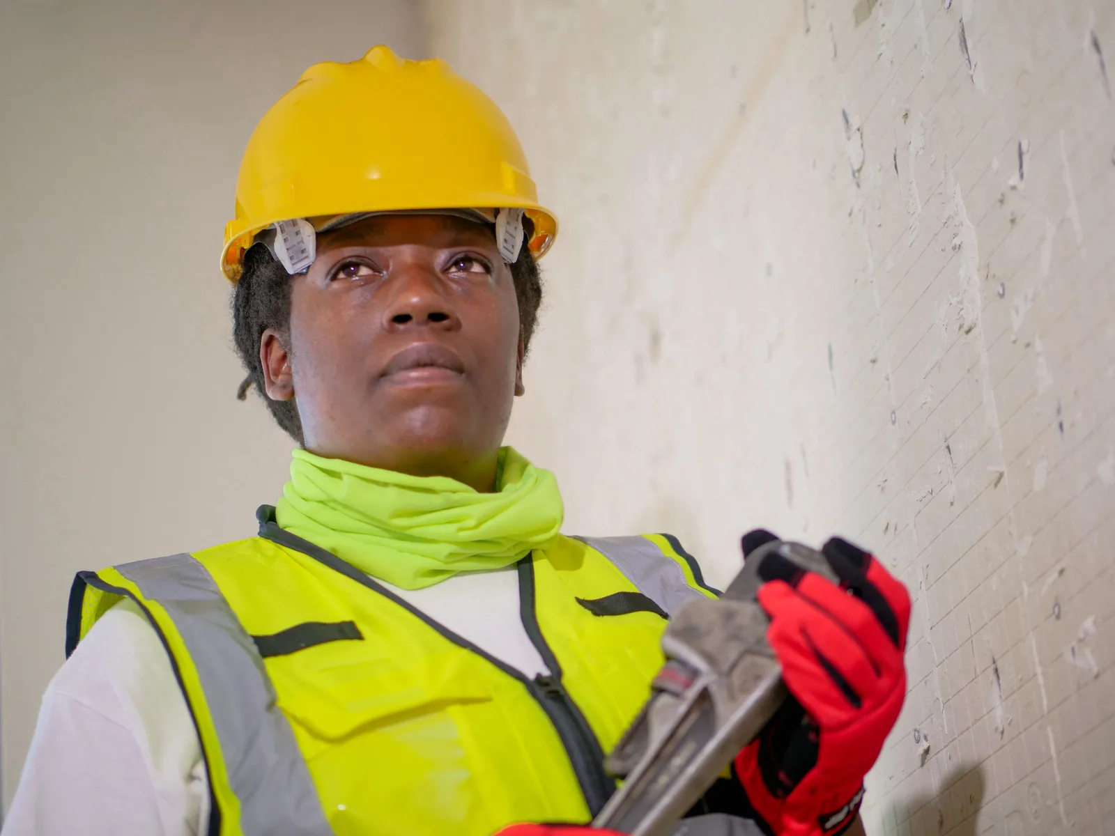 A worker in a yellow hard hat and high visibility vest holding a pipe wrench against a bare interior wall