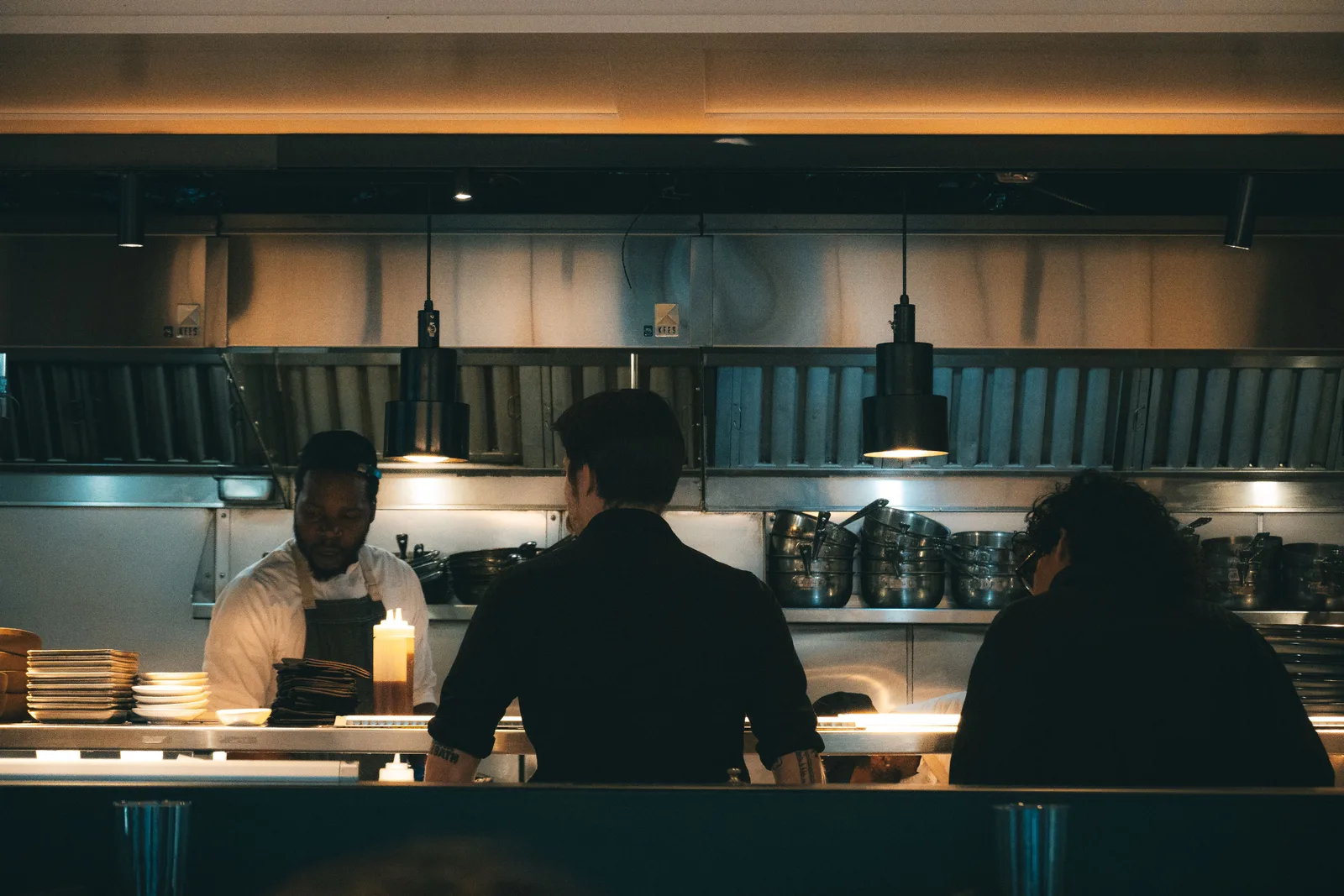 Three cooks working behind a stainless steel pass under heat lamps in a restaurant kitchen at night, with stacked plates and pans on the shelf behind them