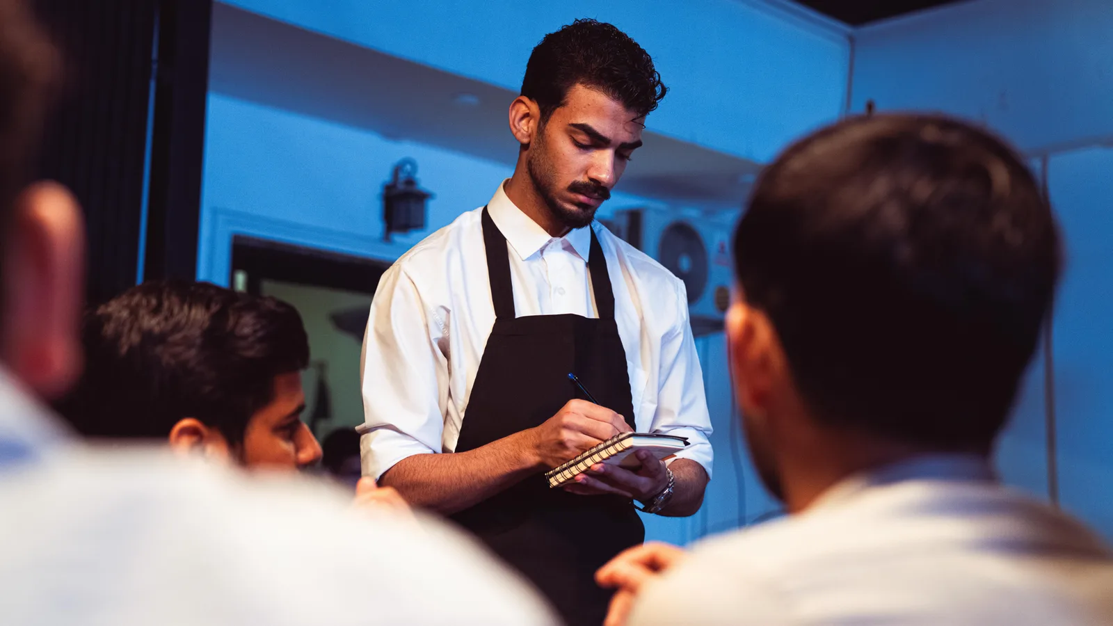 A waiter in a white shirt and black apron writing an order on a notepad at a table of seated diners