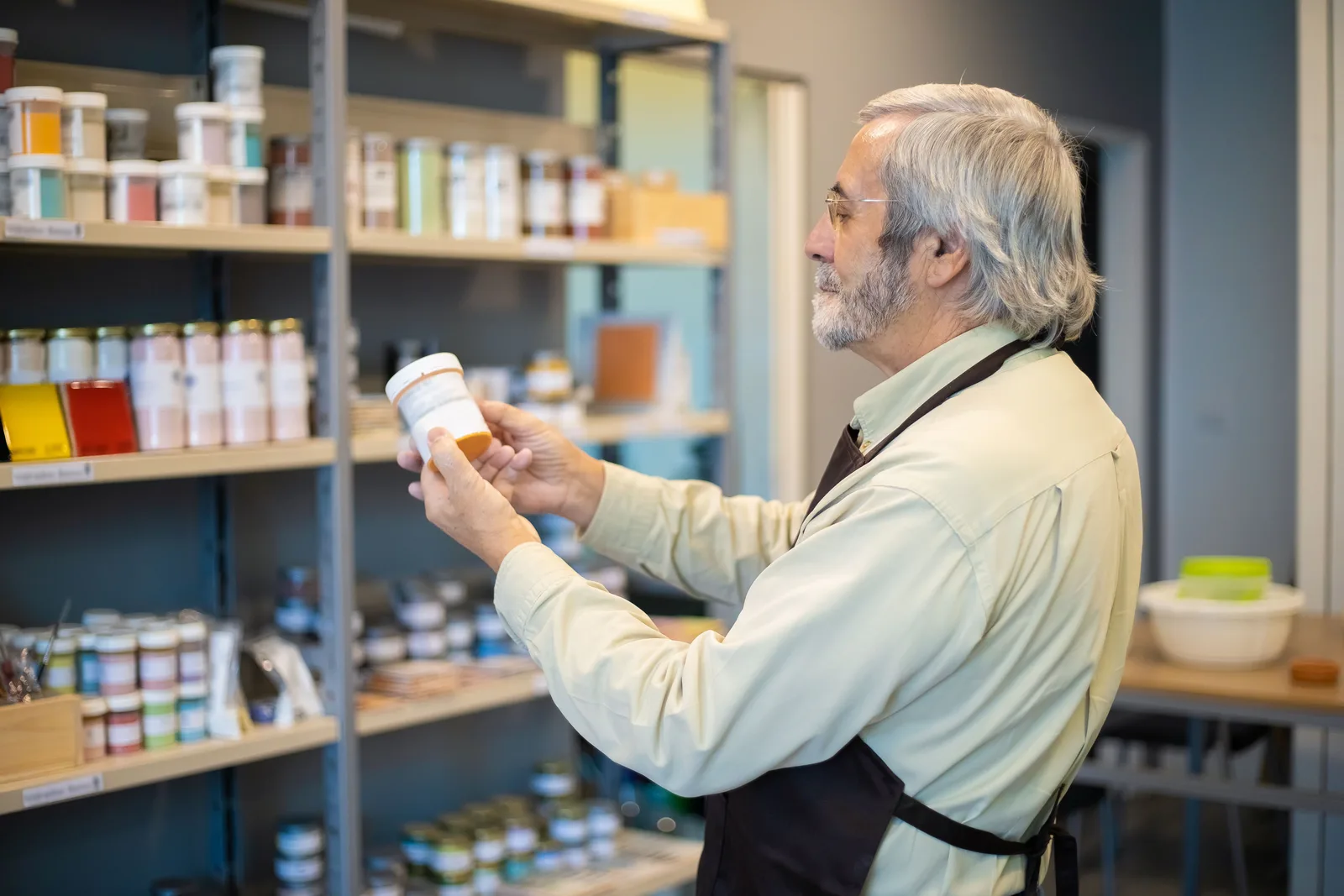 Shopkeeper in an apron checking a jar of stock against a full shop shelf