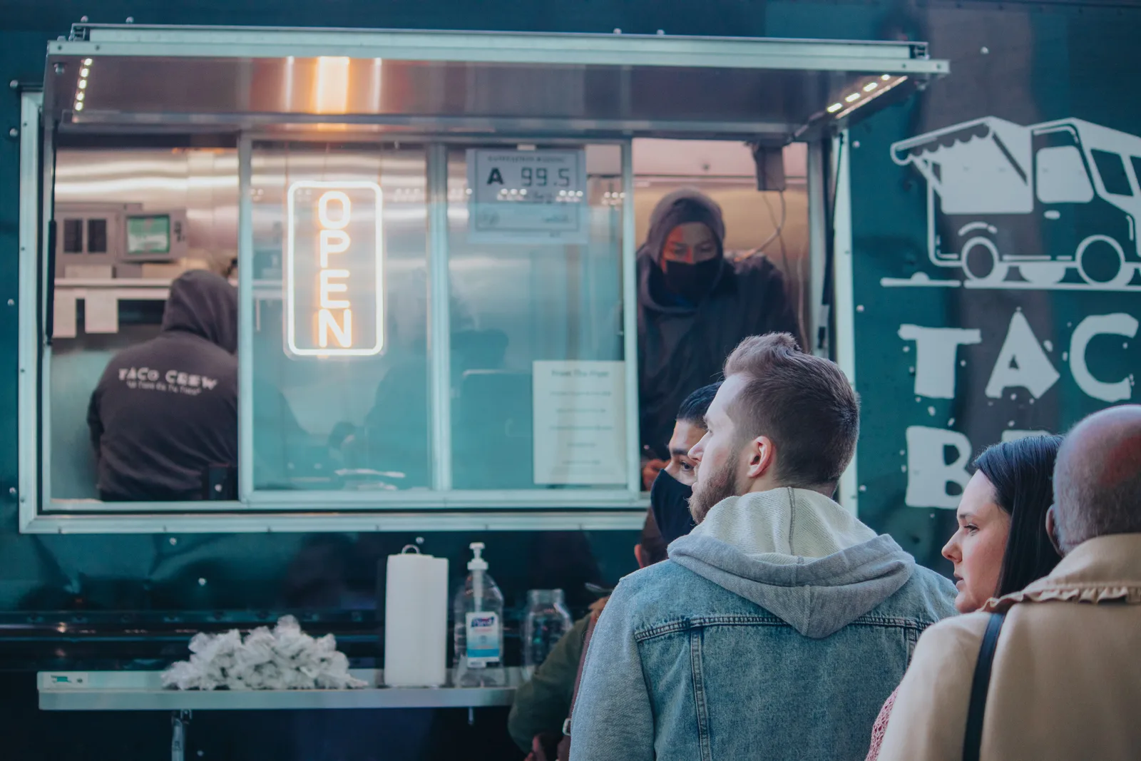 Customers queue at the lit service window of a taco food truck in the evening.