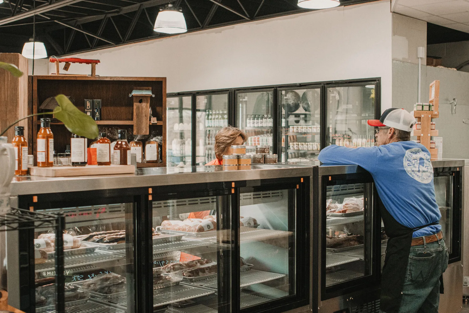 A butcher counter with refrigerated cases of packaged meat, a customer leaning on the counter and a staff member working behind it
