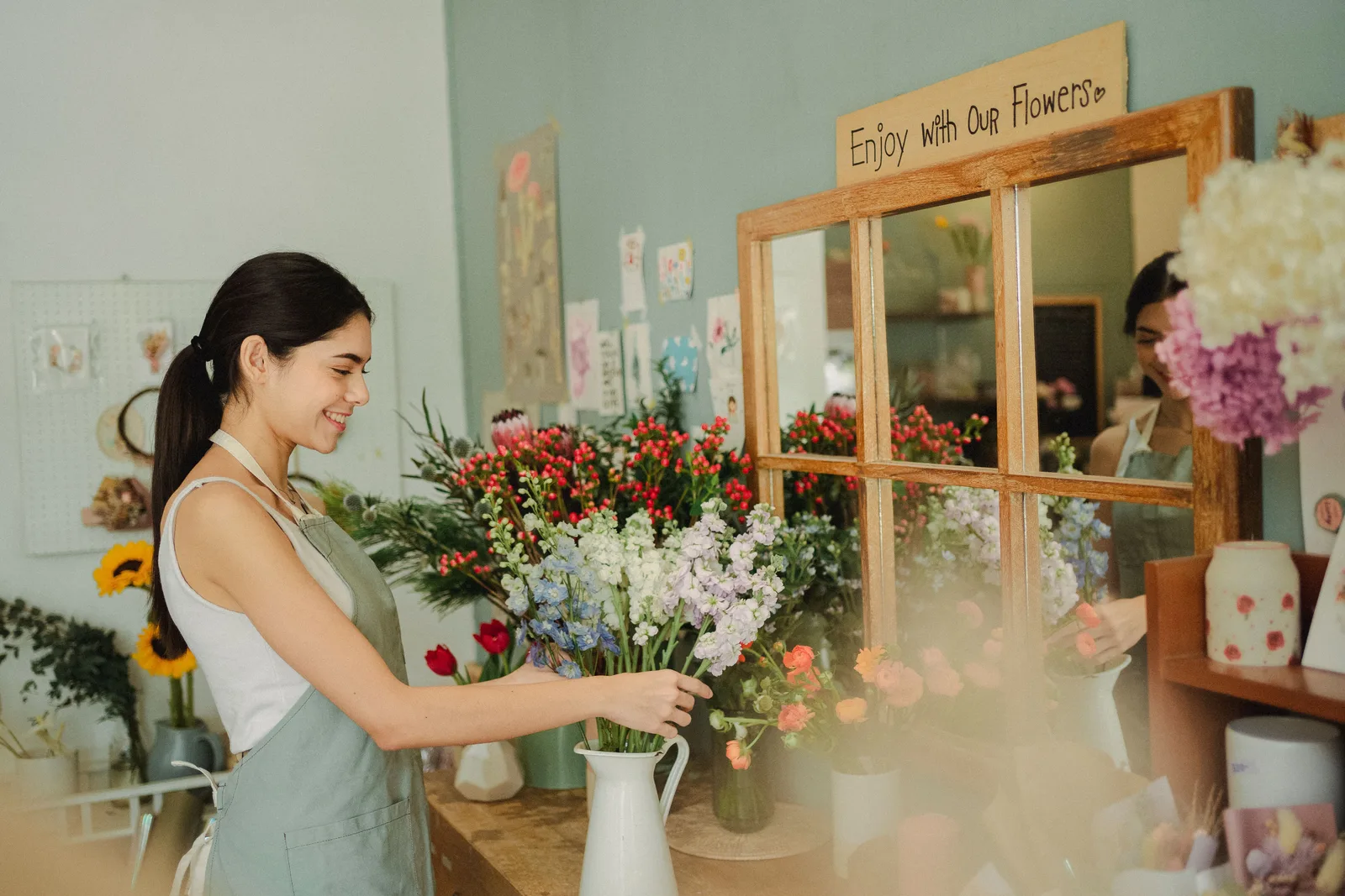 A florist arranges cut stems into a white jug at the workbench of a flower shop.
