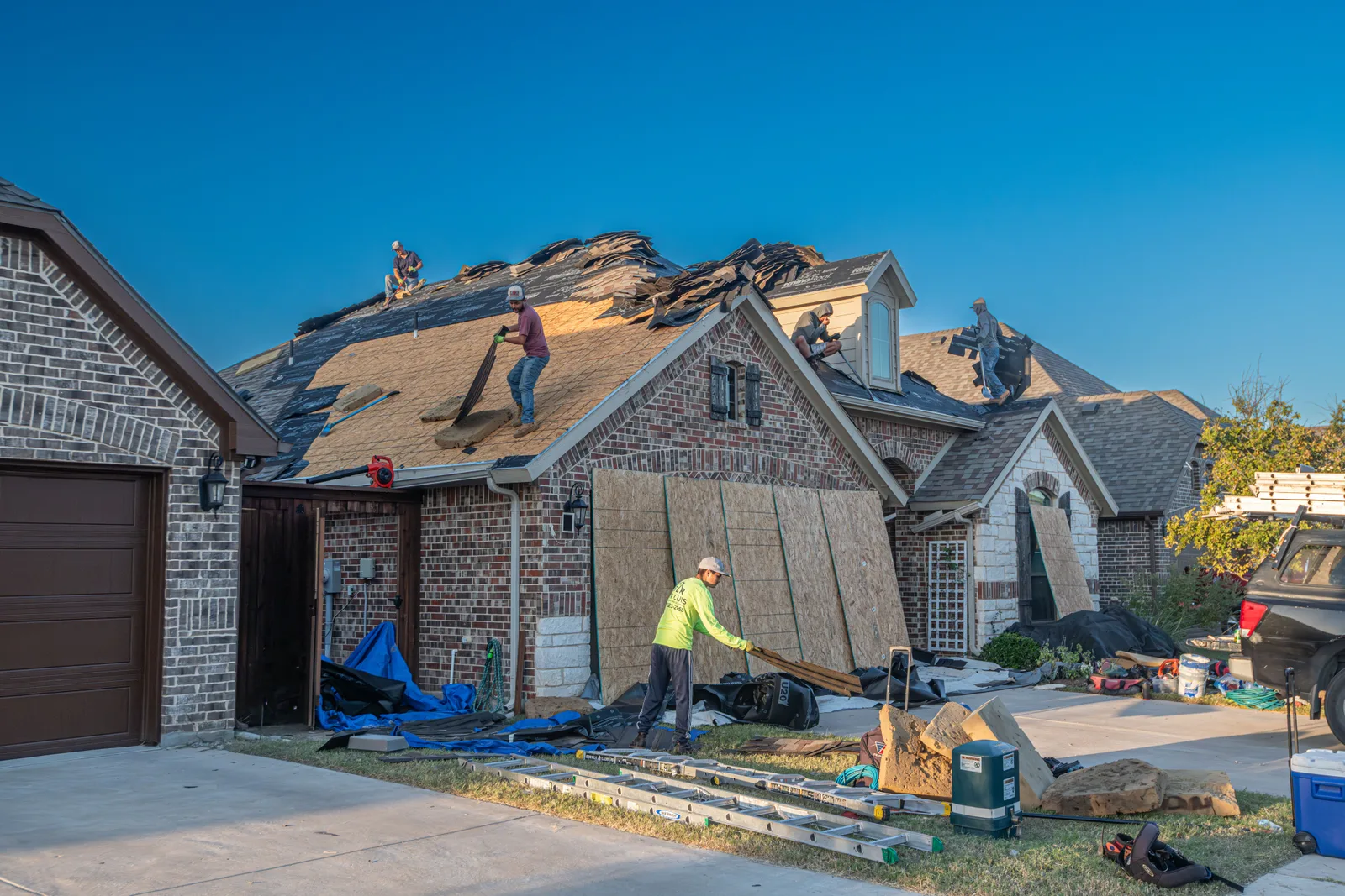 A crew working across the roof of a two storey brick house during a full roof replacement, with materials stacked on the slope