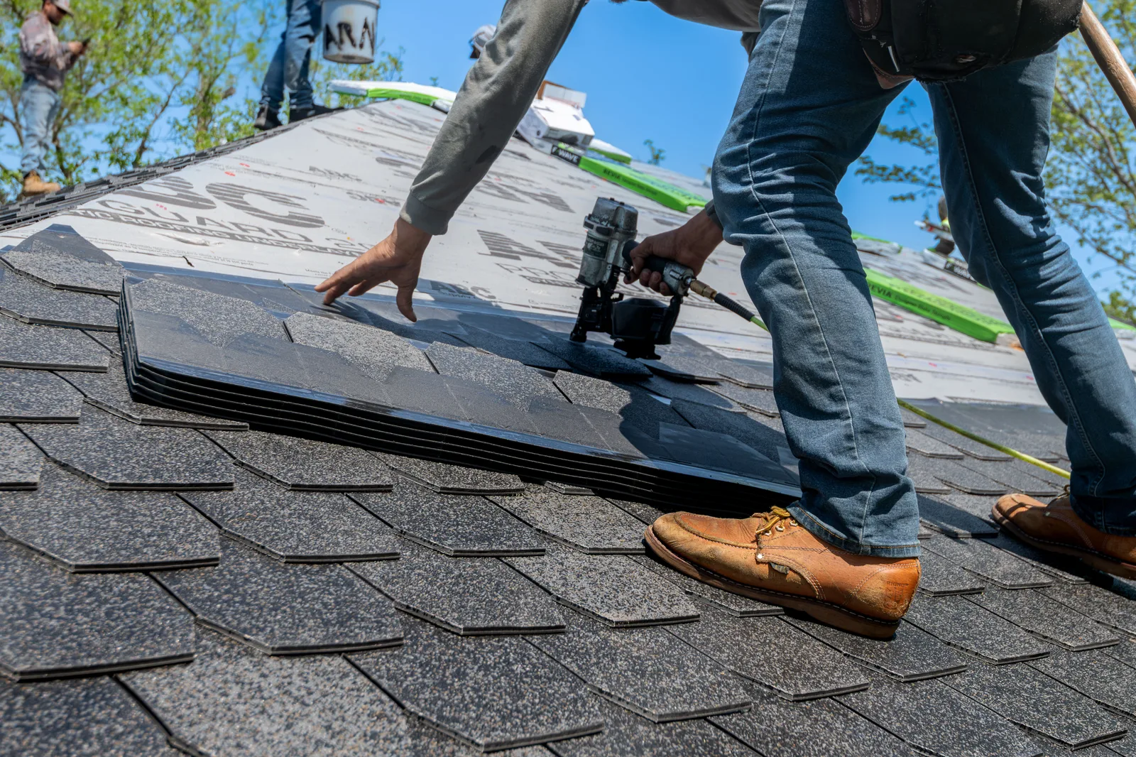 A roofer kneeling on a pitched roof, driving nails into a course of asphalt shingles with a nail gun