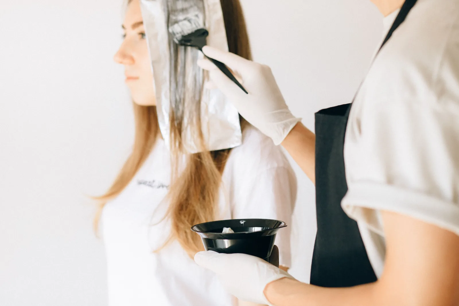 Gloved stylist applying color to a client’s hair with a tint brush and bowl