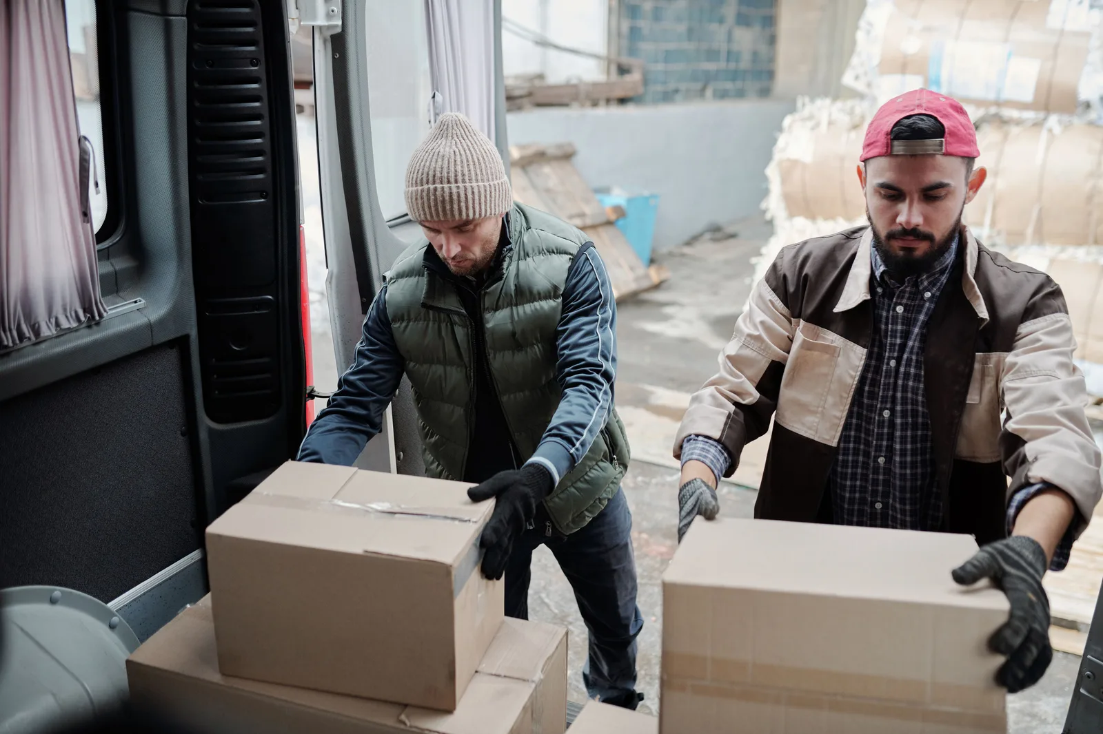 Two workers in gloves lifting cardboard cartons into the side door of a delivery van in a yard