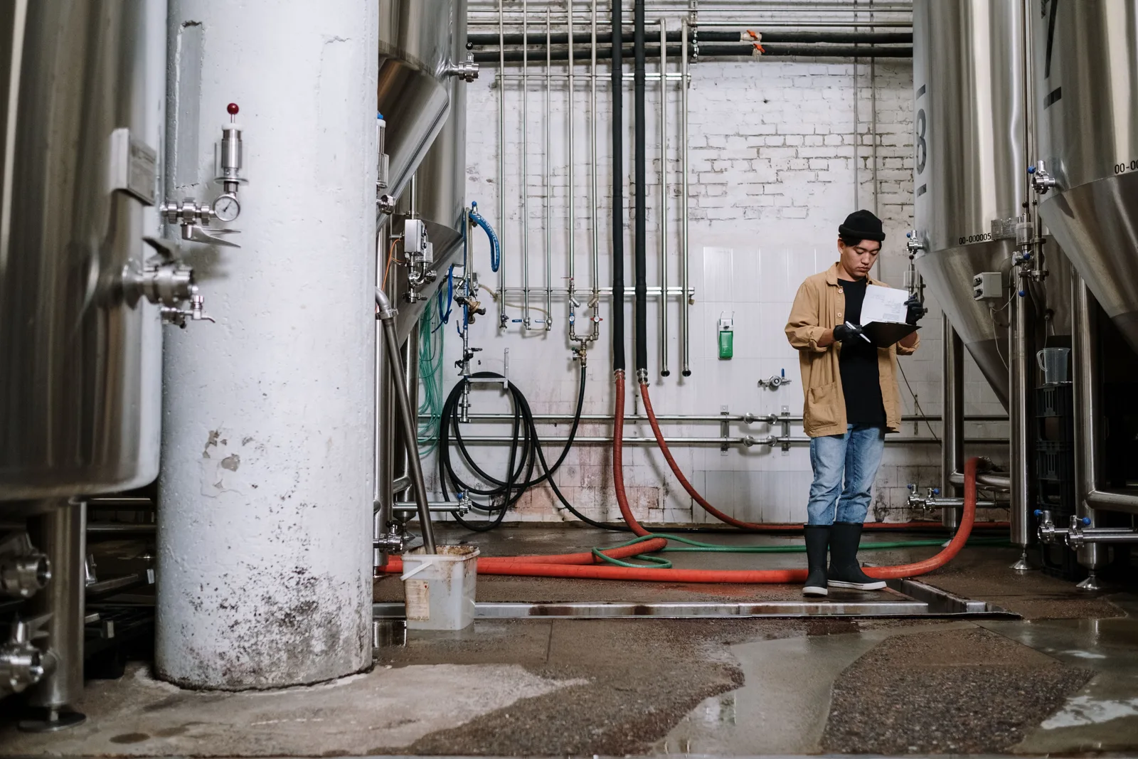 A brewery worker reading notes on a clipboard while standing between stainless steel fermentation tanks