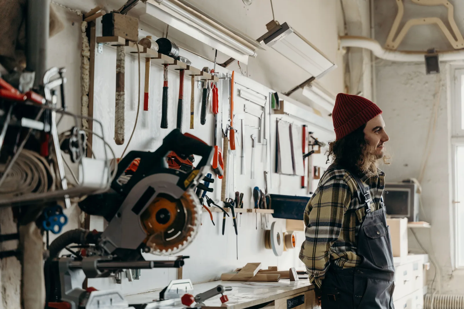 A woodworker standing beside a mitre saw in a workshop with hand tools hung on the wall behind