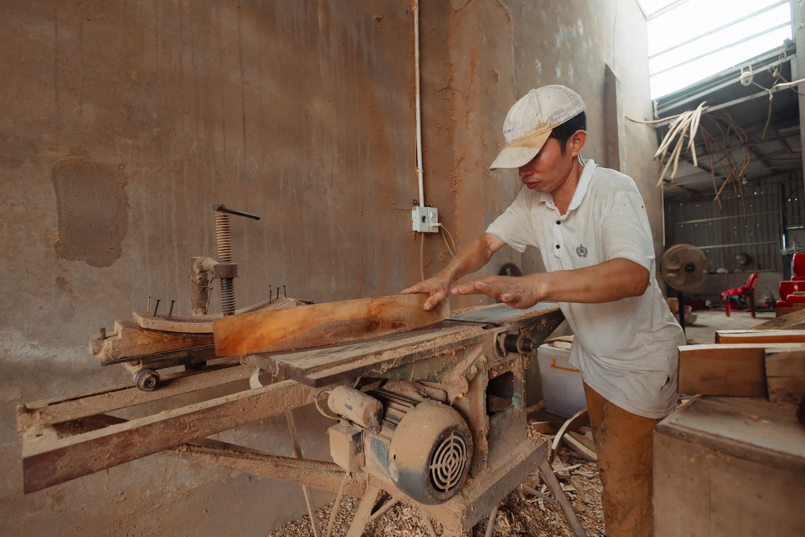 A man in a cap feeding a plank of timber across a jointer in a dusty woodworking workshop, with sawdust on the floor and offcuts stacked behind him