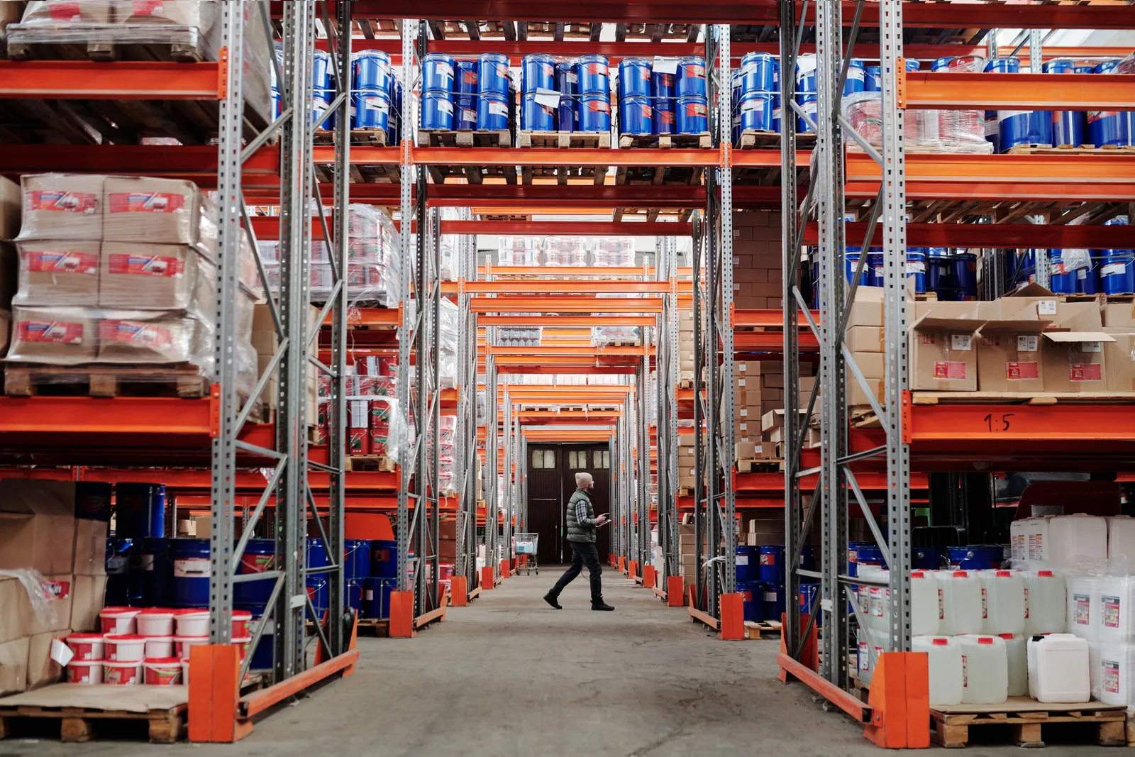 A worker walking down an aisle of tall orange pallet racking stacked with drums, cartons and plastic containers in a distribution warehouse