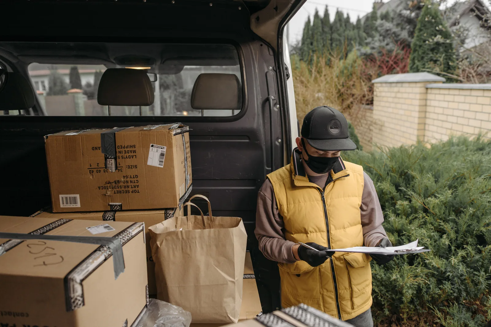 A driver in work clothes checking a printed docket on a clipboard beside a van loaded with boxes and paper bags.