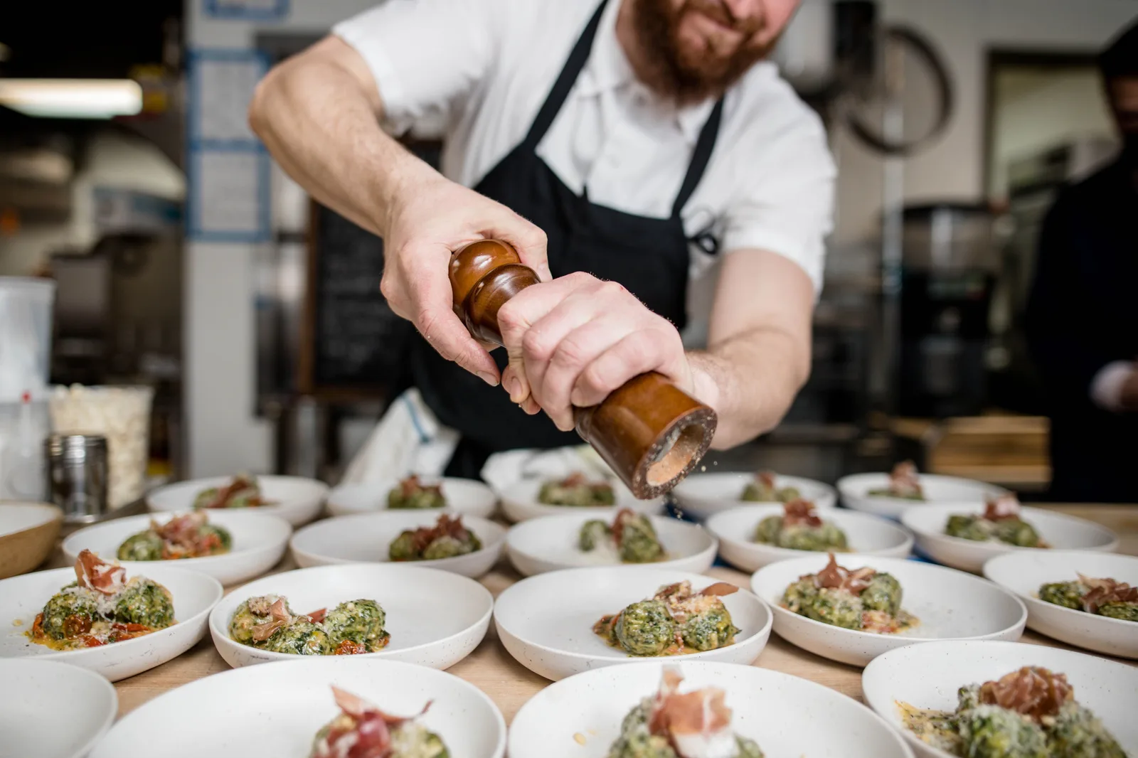 A chef in a black apron grinding pepper over a long row of plated dishes on a kitchen pass