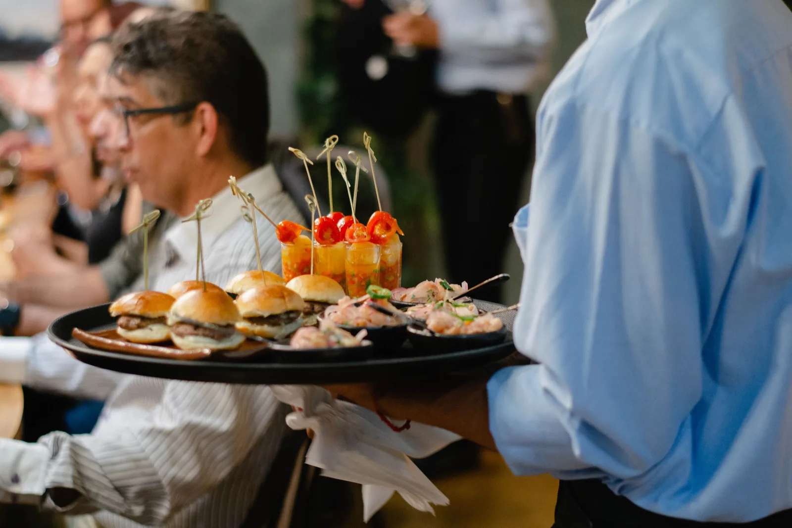 A server in a blue shirt carrying a tray of sliders and small plates through a crowded dining room