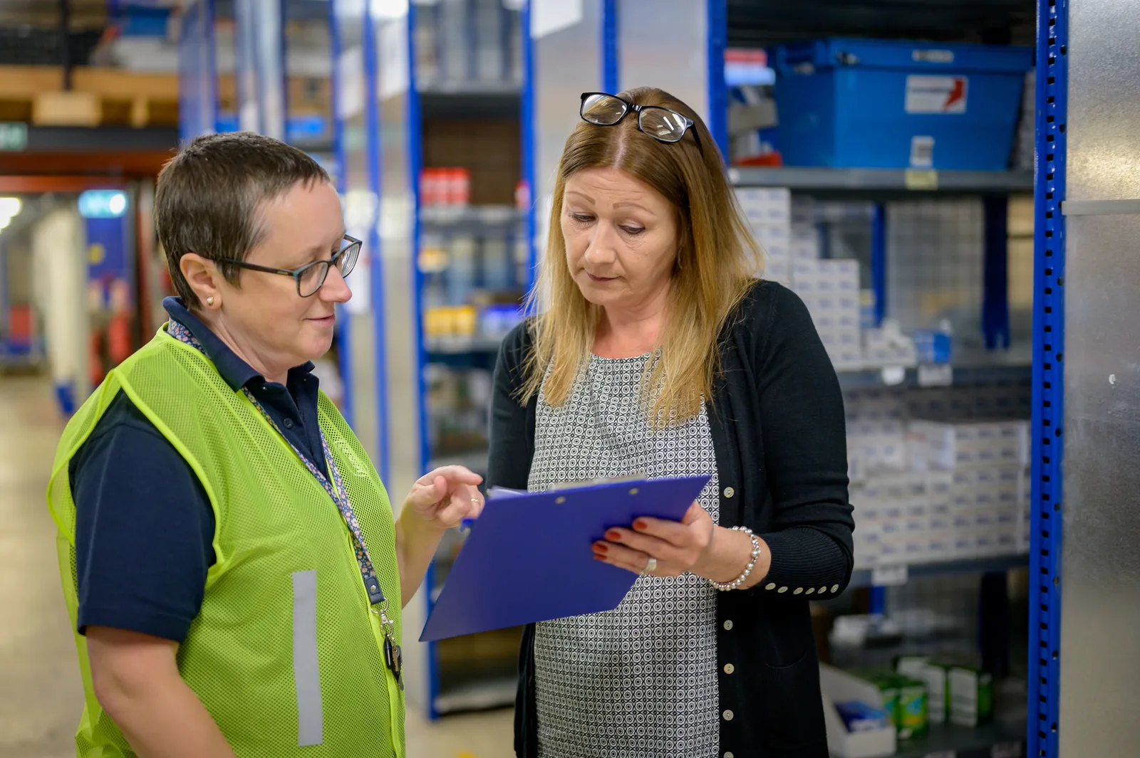 Two workers standing beside metal warehouse shelving, reviewing a checklist on a clipboard together