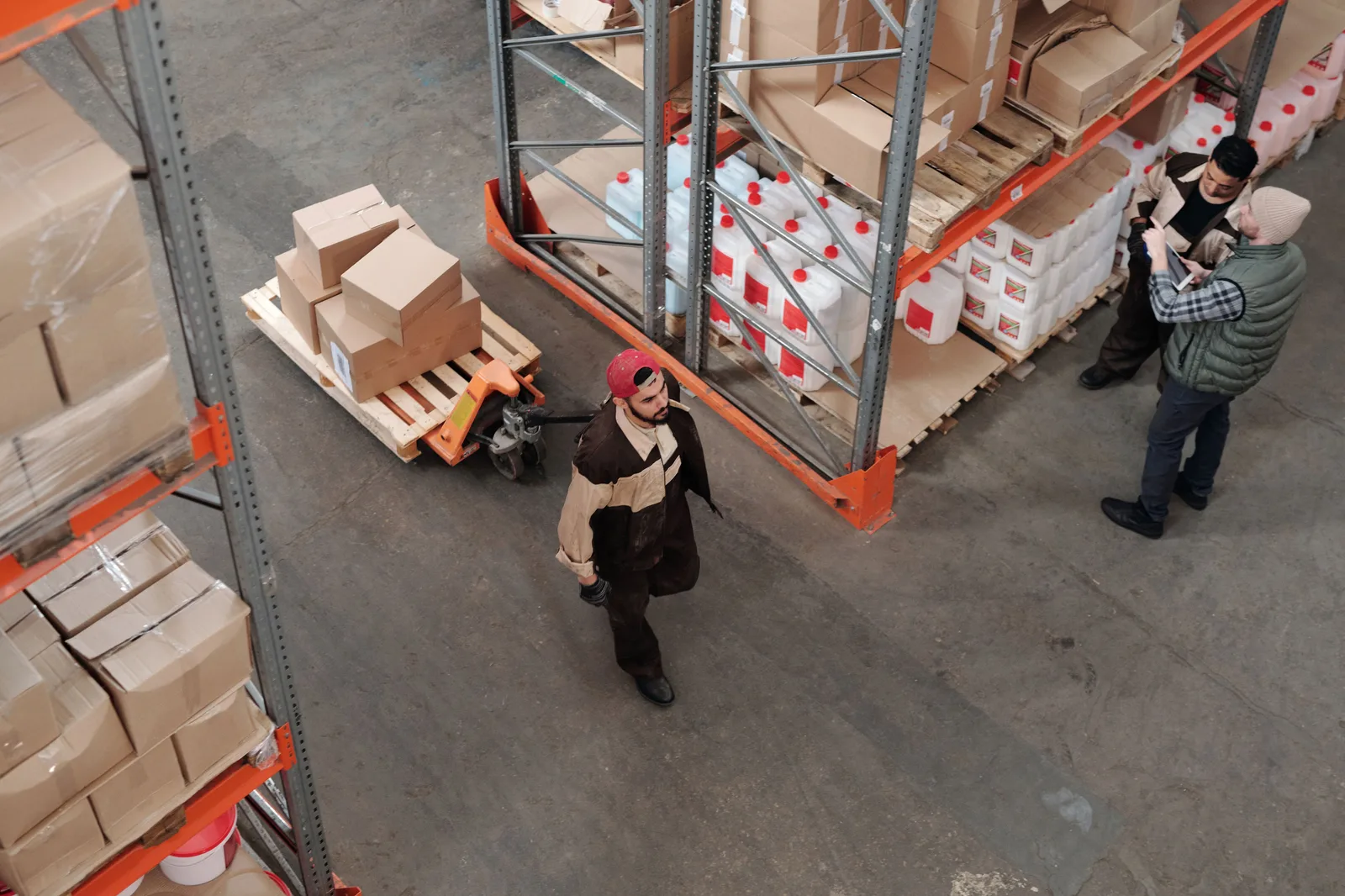 Overhead view of a warehouse aisle where one worker moves a loaded pallet with a pallet truck while two others check stock on a tablet