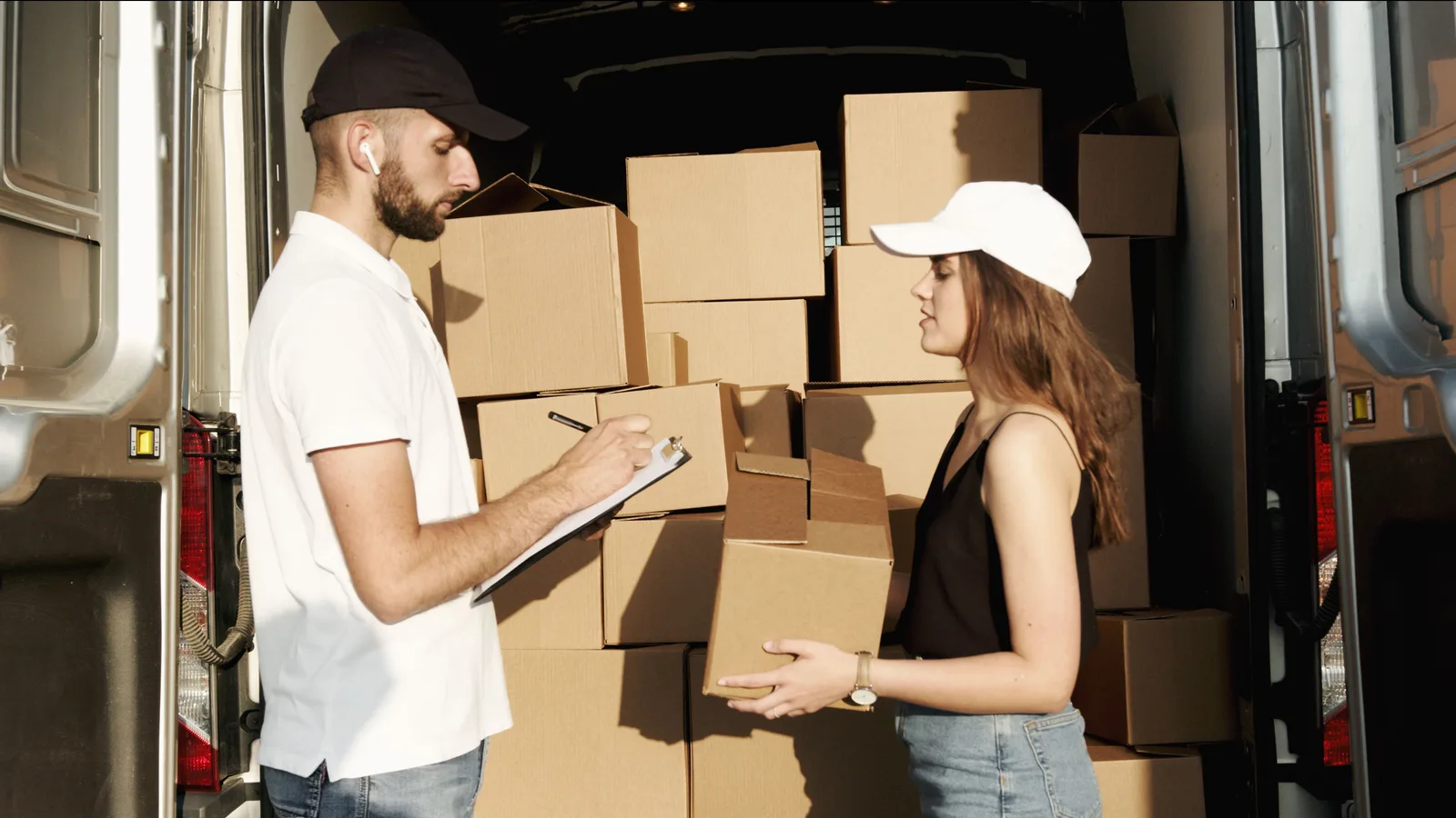 Two workers at the open back of a loaded delivery van, one signing a clipboard while the other holds a cardboard carton