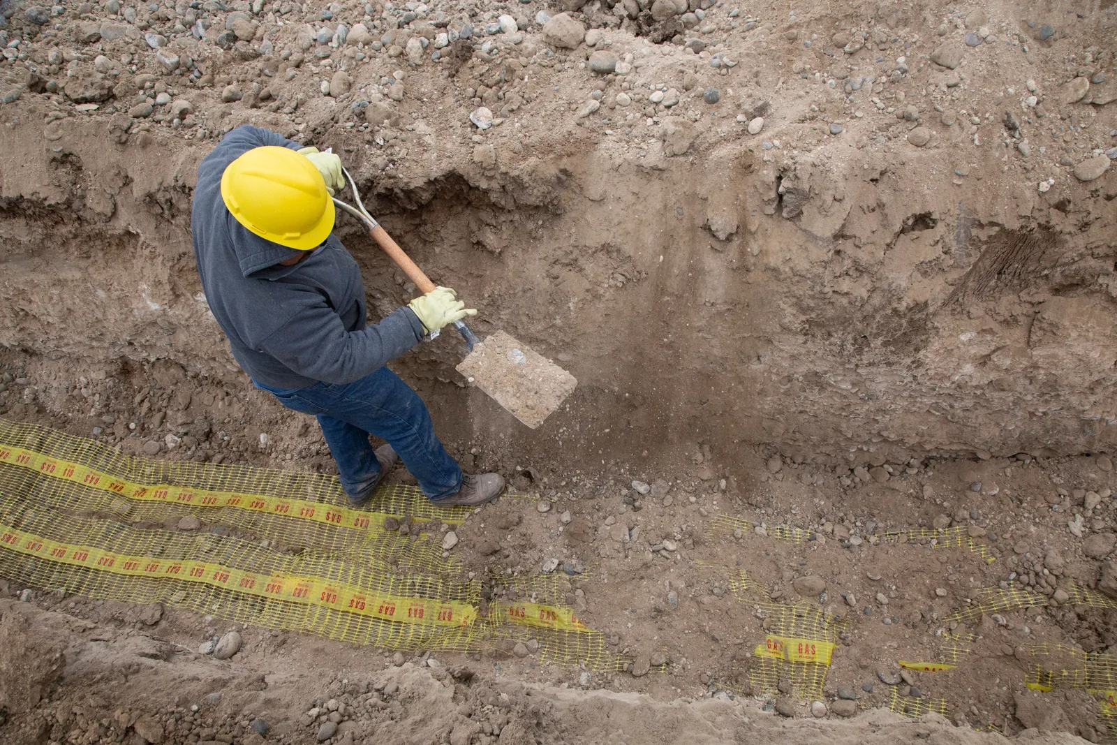 A worker in a hard hat digging a trench by hand above a marked underground utility line