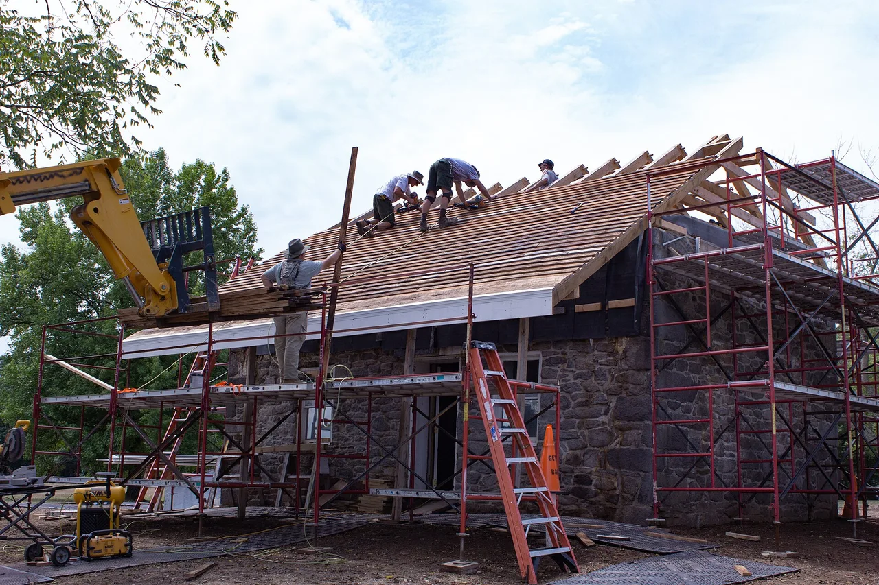 Roofing crew fastening battens across a new roof deck with a telehandler and scaffolding in place
