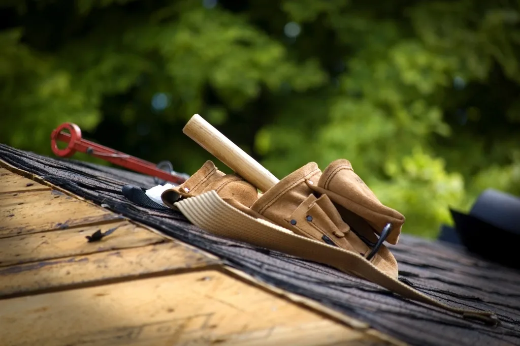 Roofer tool belt, hammer and shingle bar resting on a roof deck beside underlayment and shingles