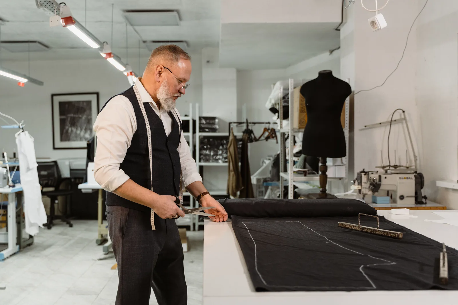 A tailor cutting a length of dark cloth on a long cutting table in a garment workshop