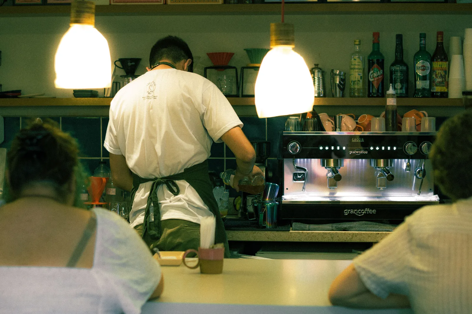 Barista working at an espresso machine behind a tiled cafe counter with customers seated at the bar