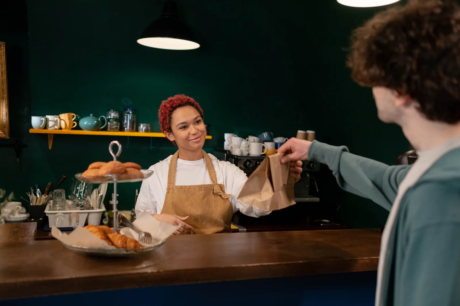 A barista hands a paper bag of pastries across the counter to a customer in a small cafe