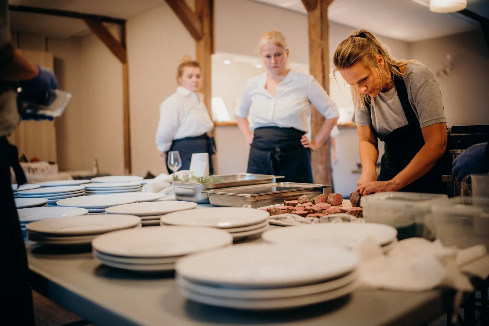 Cooks working along a stainless steel pass, plating dishes as servers wait at the end of the line.