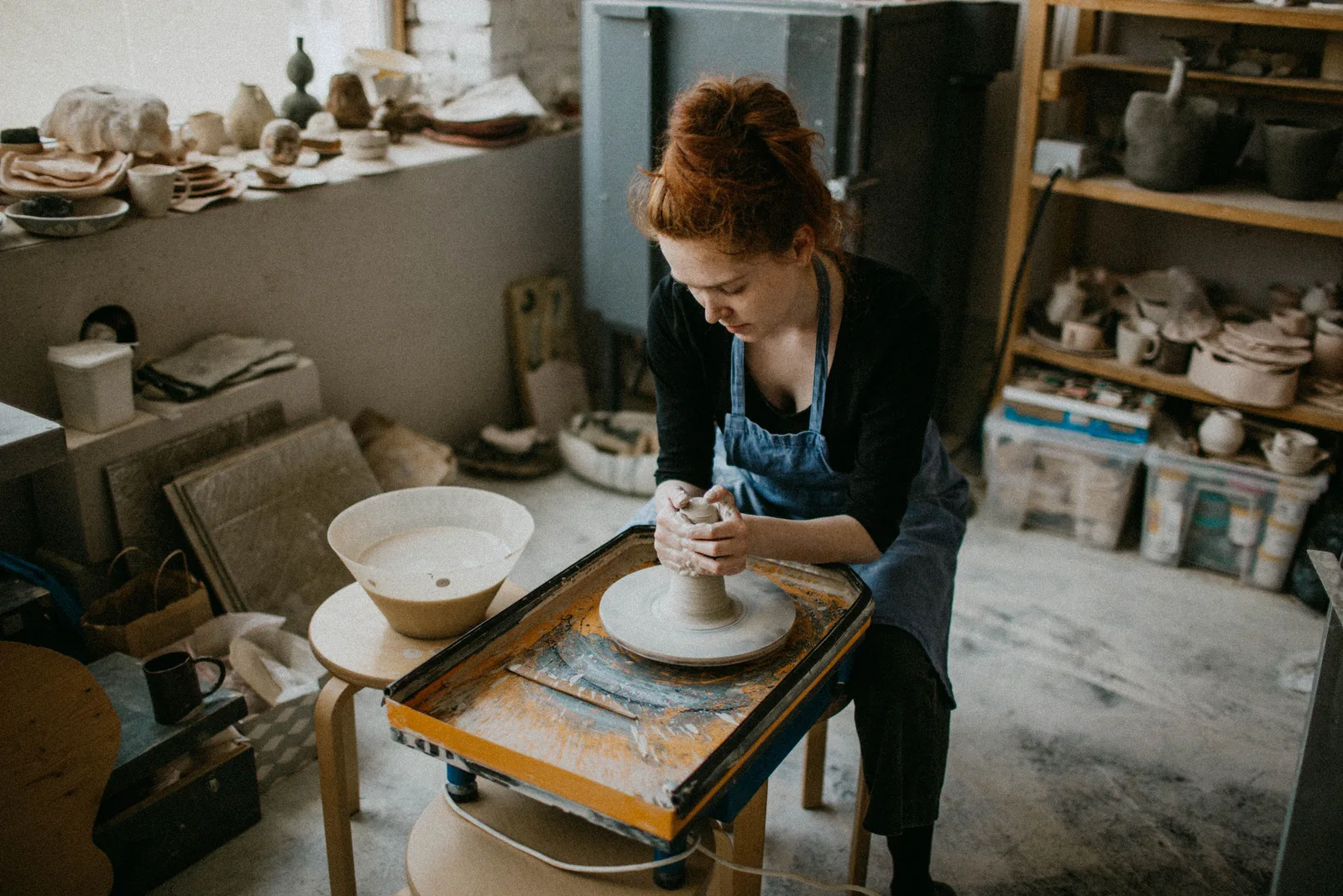 Potter shaping a bowl on the wheel in a studio lined with finished work.